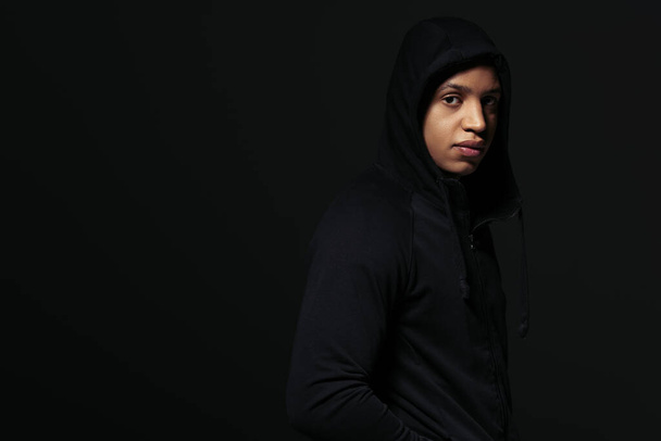 African american hooligan in hoodie looking at camera while standing isolated on black  - Valokuva, kuva