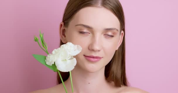 Virág érintése egy gyönyörű lány arcát természetes smink és tiszta bőr. Közelkép egy fiatal nőről rózsaszín háttérrel. Az ökológiai kozmetikumok fogalma. - Felvétel, videó