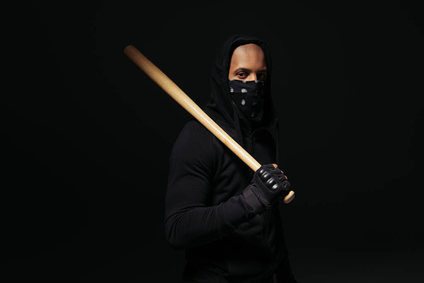 Африканский американский хулиган с маской на лице, держащий деревянную бейсбольную биту, изолированную на черном  - Фото, изображение