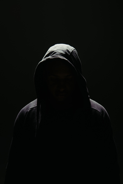 Σιλουέτα χούλιγκαν στο καπό στέκεται στο φωτισμό που απομονώνεται στο μαύρο  - Φωτογραφία, εικόνα