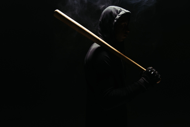 Silhouette of bandit in hoodie holding baseball bat on black background with lighting and smoke  - Фото, изображение