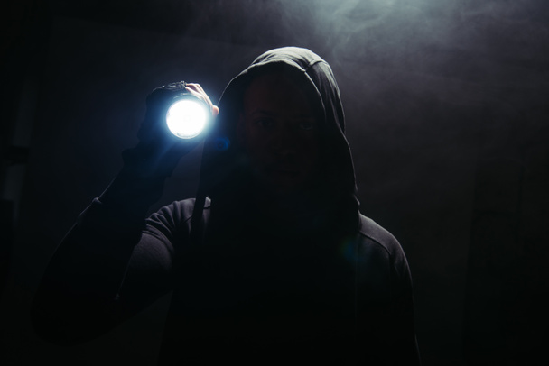Silhouette of hooligan holding flashlight on black background with smoke  - Foto, imagen