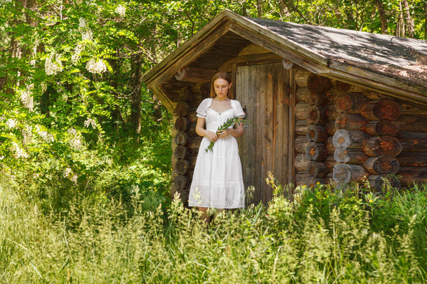 Beautiful girl in a white dress with flowers near a wooden house close-up - Foto, Bild