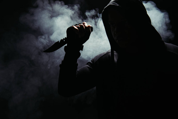 Silhouette of african american bandit in hood holding knife on black background with smoke  - 写真・画像