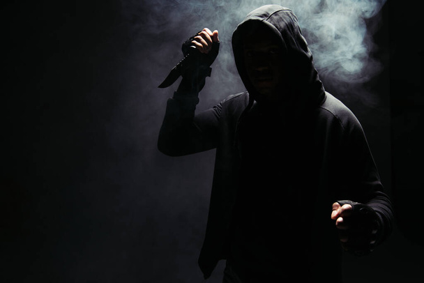 Silhouette of african american bandit in hoodie holding knife on black background with smoke  - Zdjęcie, obraz