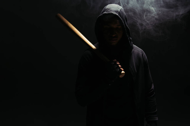 Silhouette of african american hooligan in hood holding baseball bat on black background with smoke  - Photo, Image
