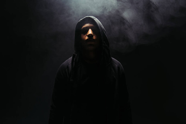 African american hooligan in hoodie standing on black background with smoke  - Photo, image
