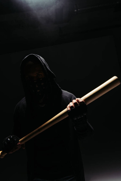 Silhouette of african american vandal holding wooden baseball bat on black background with smoke  - Фото, зображення