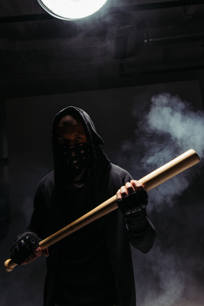 Silhouette of african american vandal with mask on face holding baseball bat on black background with smoke  - Foto, Imagem
