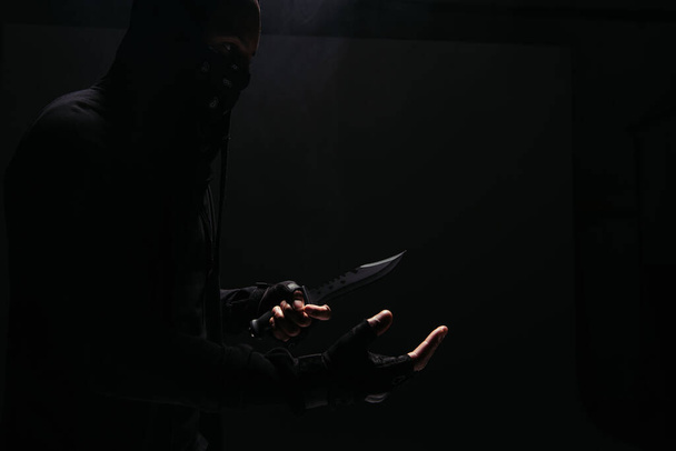 Бічний вид на афроамериканського бандита в масці, який тримає ніж ізольований на чорному  - Фото, зображення
