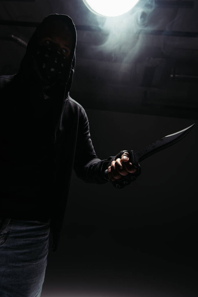 Silhouette of african american hooligan with obscure face holding knife on black background with smoke  - 写真・画像