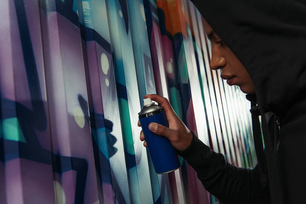 Side view of african american hooligan in hoodie drawing graffiti on wall on urban street  - Zdjęcie, obraz