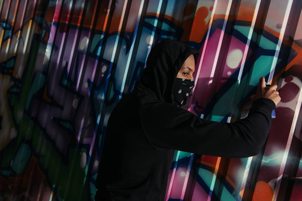 Vista lateral del vándalo afroamericano mirando hacia otro lado mientras dibuja graffiti en la pared al aire libre por la noche  - Foto, Imagen