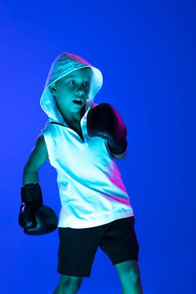 Sport and fashion. Stylish boy, beginner boxer training isolated over blue background in neon light. Concept of sport, fashion, studying, achievements, skills. Kid practicing punch. Copy space for ad - Photo, Image