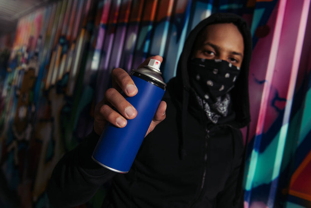 Blurred african american vandal holding spray paint near graffiti on wall  - Photo, Image
