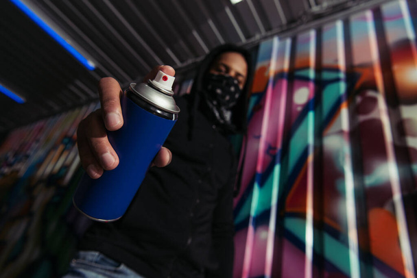 Low angle view of blurred african american vandal holding spray paint near graffiti on wall  - 写真・画像