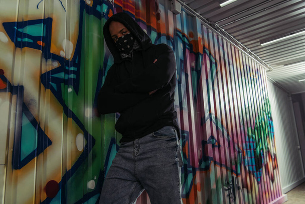 African american vandal in mask and hoodie crossing arms near graffiti on wall  - Fotó, kép