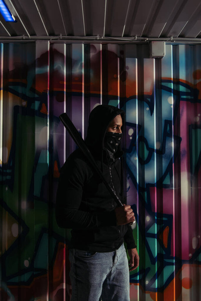 Side view of african american hooligan in mask and hoodie holding baseball bat near graffiti on wall  - Foto, immagini