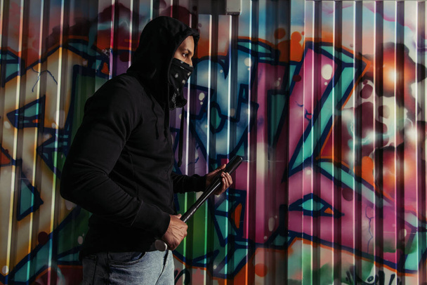 Side view of african american vandal holding baseball bat near graffiti on wall on street  - Fotó, kép
