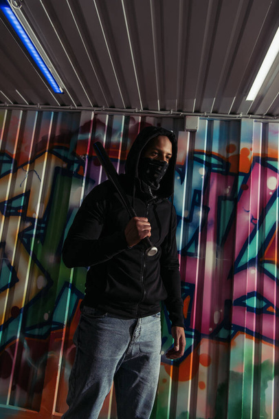 African american vandal in mask holding baseball bat near graffiti on wall  - Foto, Imagen