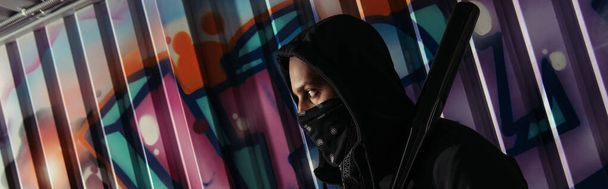 Side view of african american bandit in mask holding baseball bat near graffiti on wall, banner  - Photo, image