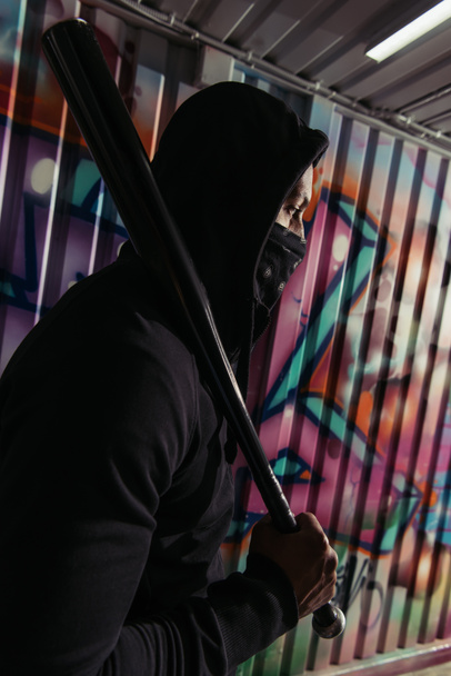 Vista lateral del vándalo afroamericano en máscara sosteniendo bate de béisbol cerca de graffiti en la pared  - Foto, Imagen