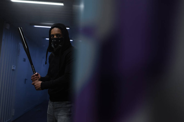 African american hooligan with mask on face holding baseball bat near blurred wall in garage - Fotó, kép
