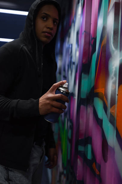 Wazig Afrikaans-Amerikaanse vandaal in capuchon schilderen graffiti en weg te kijken in de garage  - Foto, afbeelding
