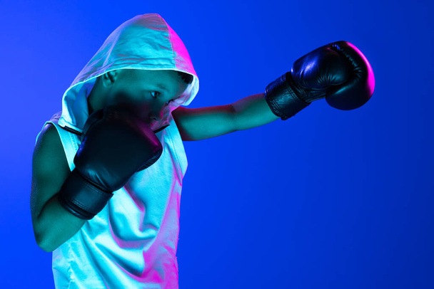 Punch. Stylish boy, beginner boxer training isolated over blue background in neon light. Concept of sport, fashion, studying, achievements, skills. Copy space for ad - Zdjęcie, obraz
