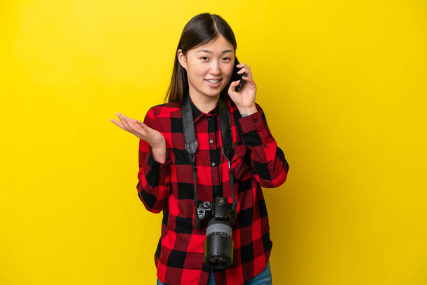 Young photographer Chinese woman isolated on yellow background keeping a conversation with the mobile phone with someone - Foto, Imagem