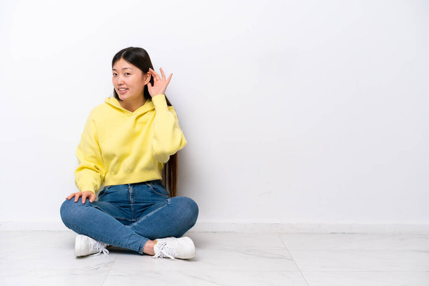 Молодая китаянка сидит на полу изолированный на белой стене, слушая что-то, положив руку на ухо - Фото, изображение
