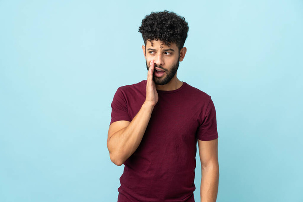 Young Moroccan man isolated on blue background whispering something with surprise gesture while looking to the side - Photo, Image
