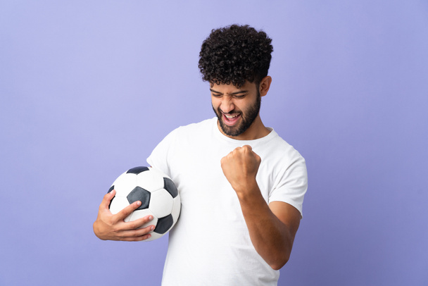 Joven hombre marroquí aislado sobre fondo púrpura con pelota de fútbol celebrando una victoria - Foto, imagen