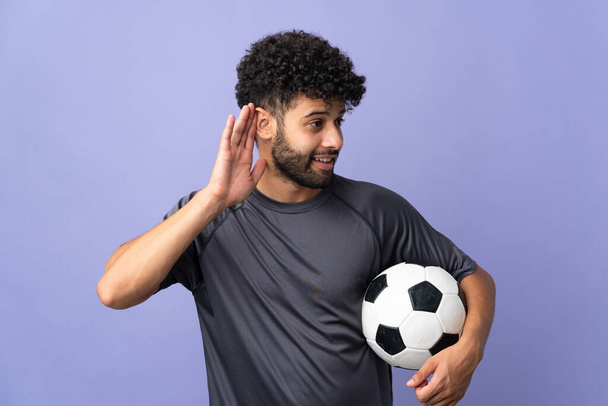 Handsome Moroccan young football player man over isolated on purple background listening to something by putting hand on the ear - Foto, Imagen