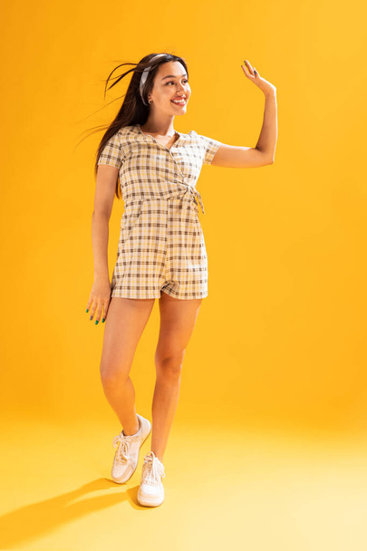 Portrait of beautiful young girl in stylish summer outfit posing, waving isolated over yellow studio background. Concept of emotions, facial expression, lifestyle, fashion, youth culture - Photo, Image