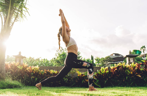 Full body of young blond female in sportswear and with braid standing in High Lunge pose during yoga practice on exotic meadow at sunrise - Foto, Imagem