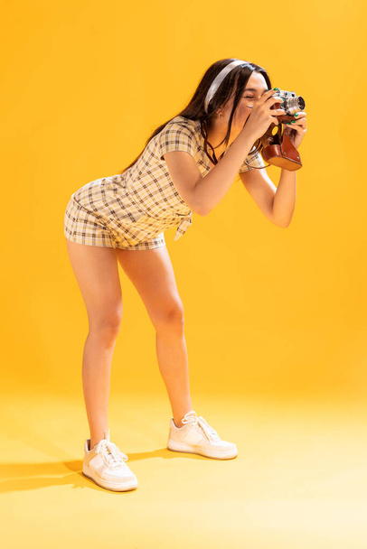 Portrait of beautiful stylish girl taking photo with vintage camera isolated over yellow studio background. Cheerful shooting. Concept of emotions, facial expression, lifestyle, fashion, youth culture - Фото, изображение