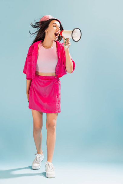 Portrait of beautiful young girl in pink panama posing, shouting in megaphone isolated over light blue studio background. Concept of emotions, facial expression, lifestyle, fashion, youth culture - 写真・画像