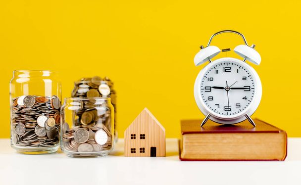 ahorros, monedas, pequeña casa de madera, despertador blanco y libro colocado en la mesa blanca Ideas de ahorro para contabilidad y finanzas - Foto, Imagen