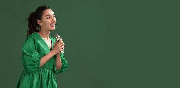 Portrait of beautiful young girl posing in dress, singing in microphone isolated over green studio background. Concept of emotions, facial expression, lifestyle, fashion, youth culture. Flyer - Valokuva, kuva