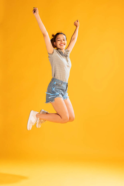 Portrait of beautiful young girl in casual outfit jumping, posing isolated over yellow studio background. Enjoyment. Concept of emotions, facial expression, lifestyle, fashion, youth culture - Photo, Image