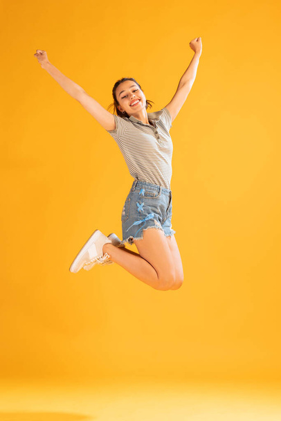 Portrait of beautiful young girl in casual outfit jumping, posing isolated over yellow background. Looks happy and delightful. Concept of emotions, facial expression, lifestyle, fashion, youth culture - Photo, image