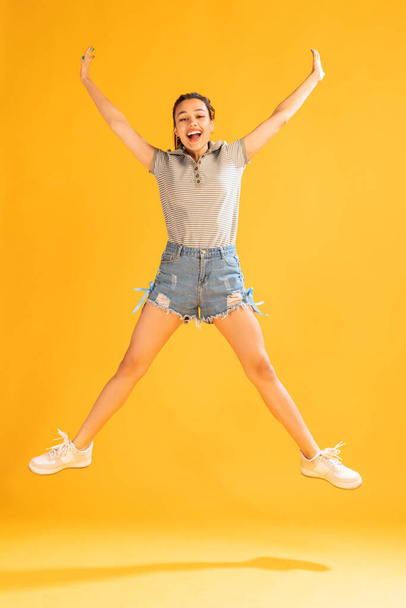 Portrait of beautiful young girl in casual outfit jumping, posing isolated over yellow studio background. Happiness. Concept of emotions, facial expression, lifestyle, fashion, youth culture - 写真・画像