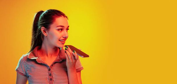 Portrait of beautiful young girl posing, recording voice message on phone isolated over yellow background in neon light. Concept of emotions, facial expression, lifestyle, fashion, youth culture - Photo, image