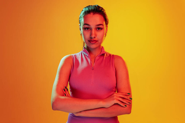 Portrait of beautiful young girl posing, attentively looking at camera isolated over yellow background in neon light. Concept of emotions, facial expression, lifestyle, fashion, youth culture - Photo, image