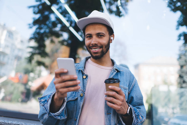 Half length portrait of cheerful male millennial smiling at camera while listening positive audio songs from media application on cellphone technology - 写真・画像
