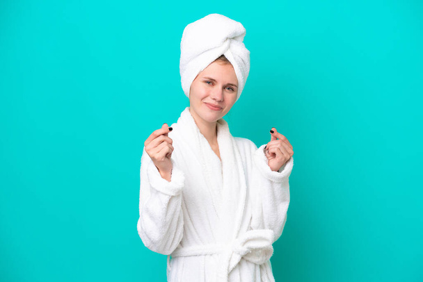 Young woman in a bathrobe isolated on blue background making money gesture - Fotoğraf, Görsel
