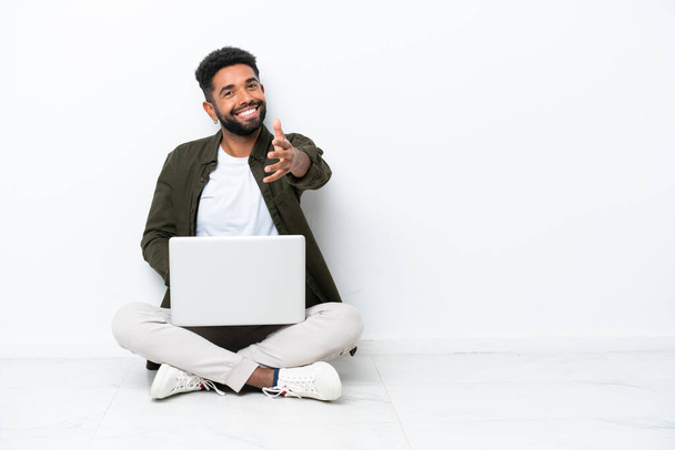 Young Brazilian man with a laptop sitting on the floor isolated on white shaking hands for closing a good deal - Fotografie, Obrázek