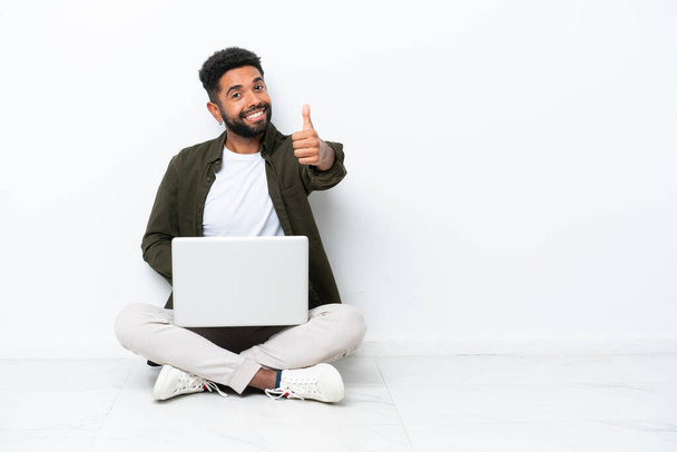 Young Brazilian man with a laptop sitting on the floor isolated on white with thumbs up because something good has happened - Photo, image