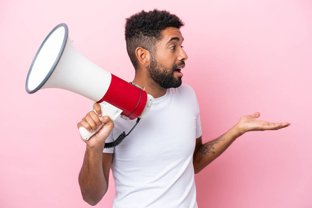 Young Brazilian man isolated on pink background holding a megaphone and with surprise facial expression - Zdjęcie, obraz